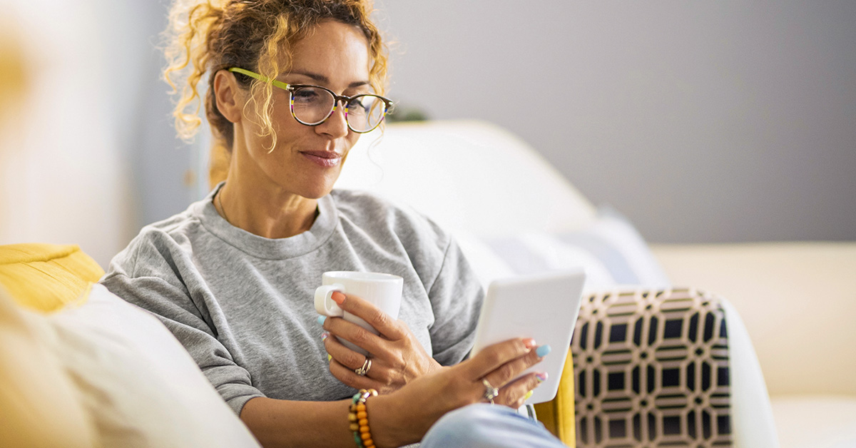 Une femme assise sur un canapé, tenant une tasse blanche et consultant des conseils de gestion budgétaire sur une tablette.