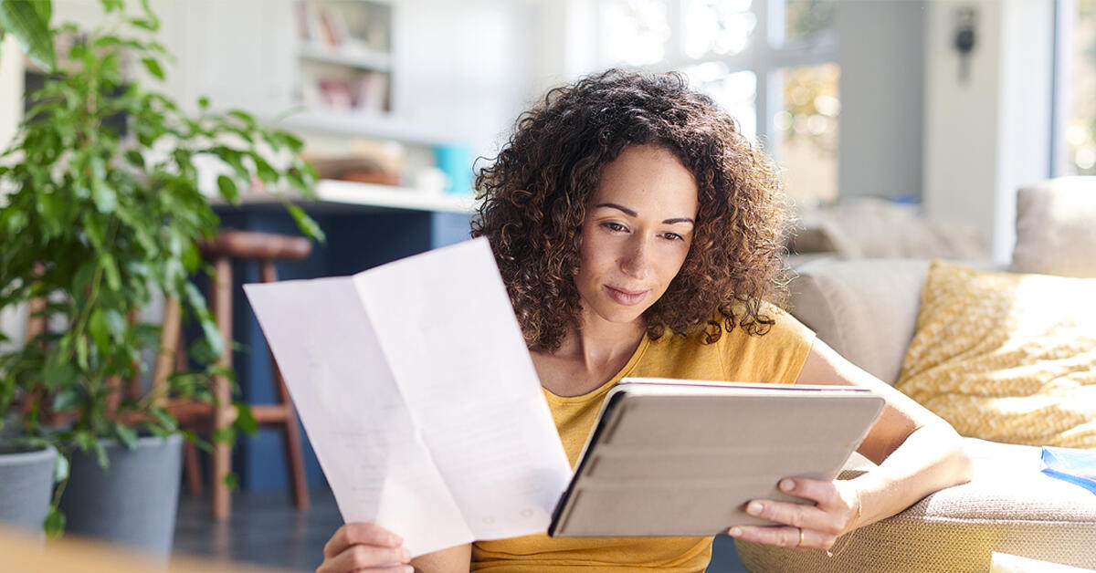 Une femme dans son salon tient un document papier dans sa main droite et regarde sa tablette qu'elle tient dans sa main gauche.