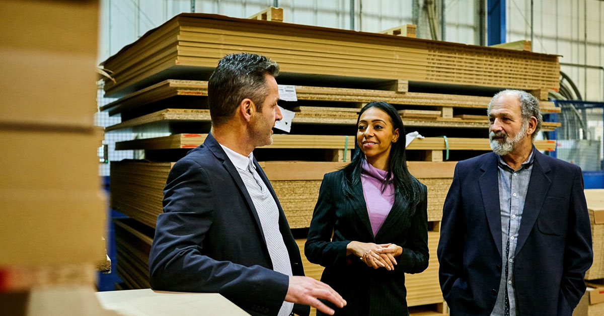 Trois personnes sont en pleine discussion dans un entrepôt industriel, au cœur d’un processus de transfert d’entreprise.]